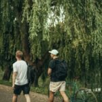 Two men with backpacks walking alongside a green bicycle under a willow tree on a park path.